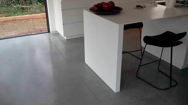 Grijze woonbetonvloer bij een wit keukeneiland en een raam naar de tuin
